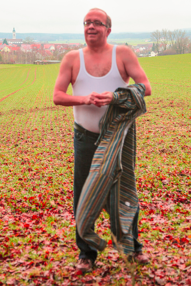 Man strips naked and undresses his shirt outdoors in the fields in front of a village.