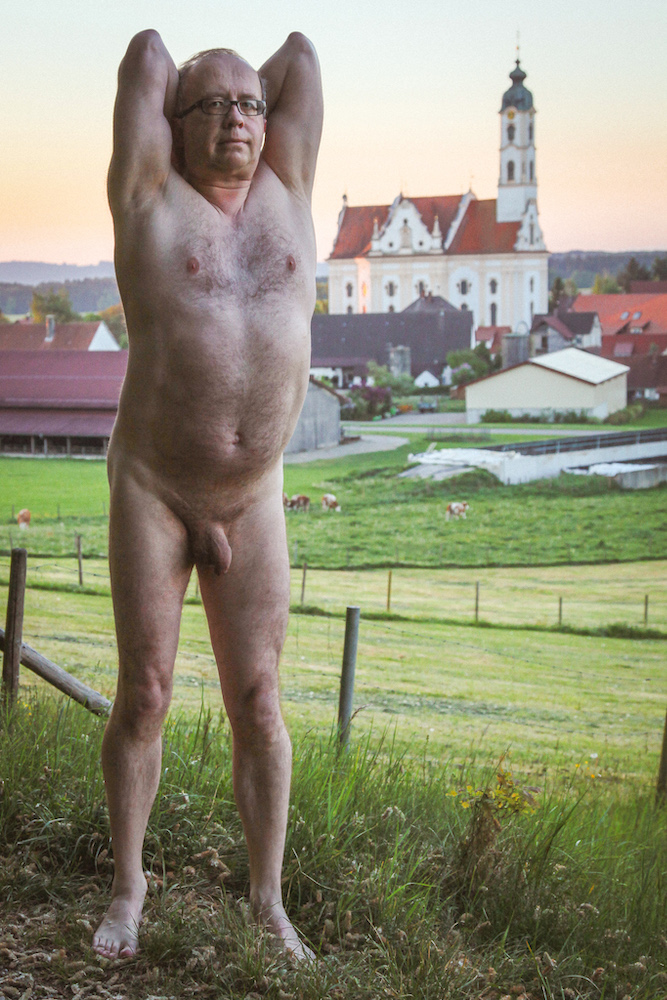 Stark naked man stretches in front of a village