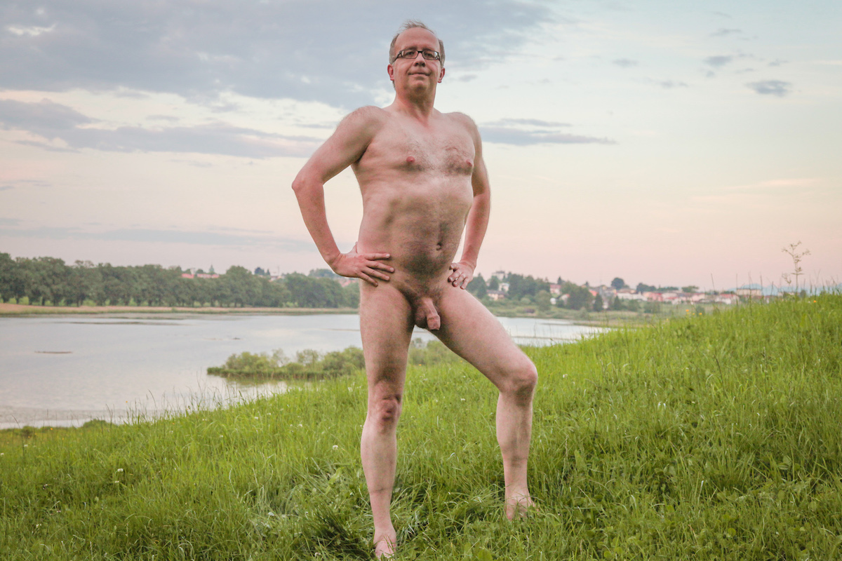 Stark naked man standing in the grass in front of a lake