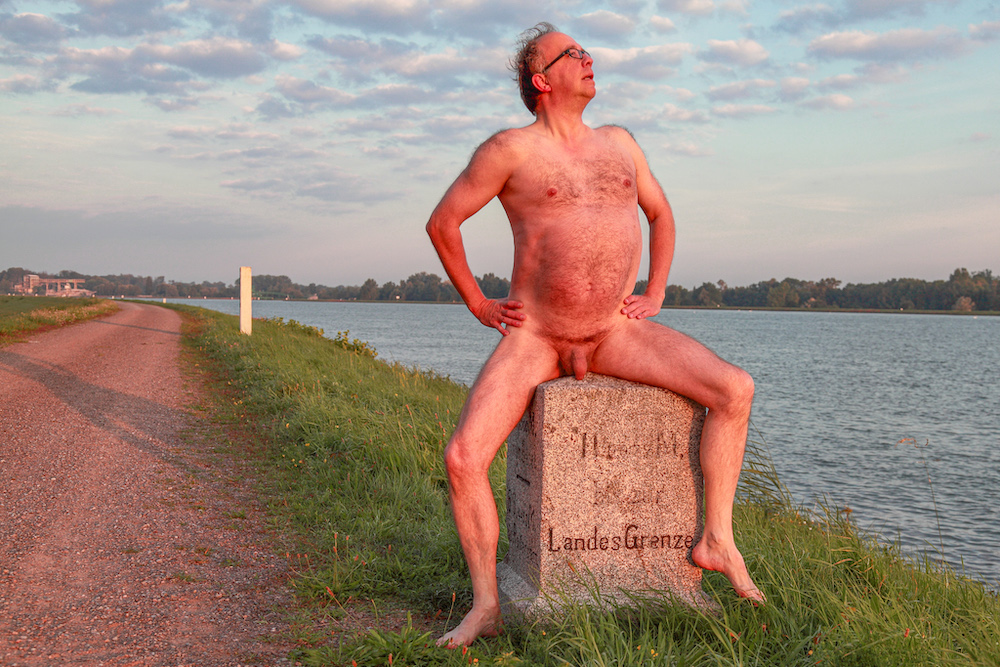 Stark naked man sitting on boundary stone on the river Rhine