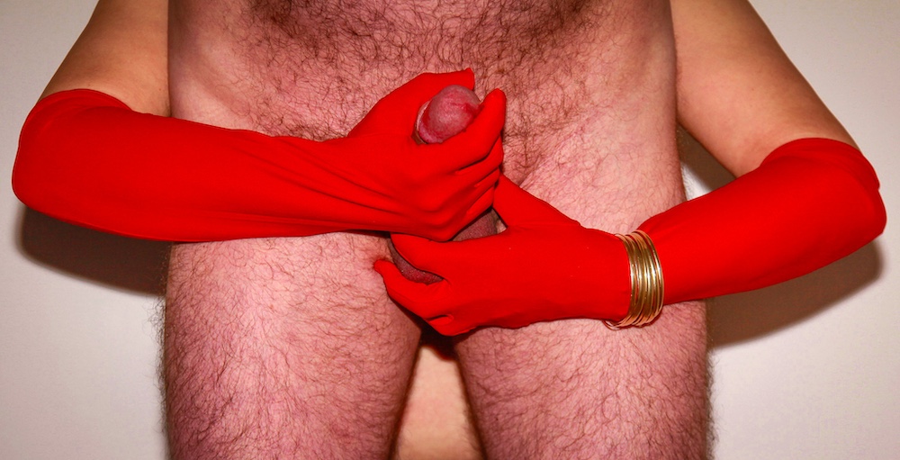 My wife holds my stiff penis shaft with one red-gloved hand so that only the glans is visible, and with the other she squeezes my testicles